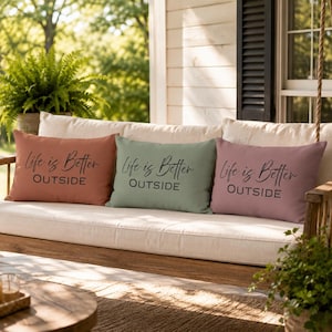Peut inclure: Trois coussins rectangulaires aux tons terracotta, vert sauge et mauve. Chaque coussin affiche la phrase "Life is Better OUTSIDE" en écriture noire. Les coussins sont disposés sur une balançoire crème.