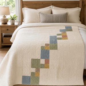 May include: A cream-coloured quilted bedspread featuring a diagonal pattern of small, multi-coloured squares in shades of muted blue, sage green, mustard yellow, and dusty rose. The quilt has a textured, grid-like stitching throughout the fabric.