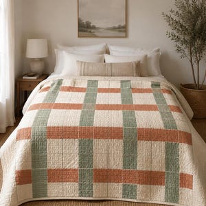 May include: A quilted bedspread featuring a large-scale plaid pattern in muted sage green and terracotta orange on an off-white background. The fabric has a visible stitched texture throughout, creating a grid design across the entire surface.