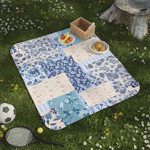 Puede incluir: Una manta de picnic con diseños florales en tonos azules y crema se extiende sobre la hierba. Una pequeña cesta, un plato de fruta y un cuenco con pasteles están sobre la manta. Un balón de fútbol y una raqueta de tenis están cerca.