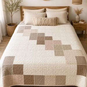 May include: A quilted bedspread featuring a grid of square patches in shades of cream, beige, taupe, and soft pink. The fabric has a textured, crinkled surface with visible stitching forming a geometric pattern across the entire surface of the quilt.