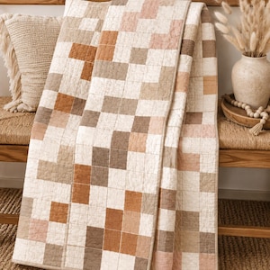 May include: A neutral-toned quilt with a geometric square pattern in shades of cream, brown, and pink. The quilt is draped over a wooden bench with a fringed pillow. The overall aesthetic is cozy and inviting, suitable for a bedroom or living room.