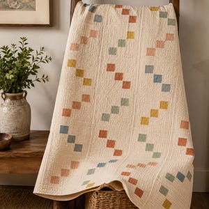 Op de afbeelding: Een cr&egrave;mekleurige quilt met een herhalend patroon van kleine, vierkante lapjes in gedempte tinten blauw, oranje, geel en groen. De stof heeft een zichtbaar getextureerd, doorgestikt oppervlak met verticale stiklijnen door het hele ontwerp.