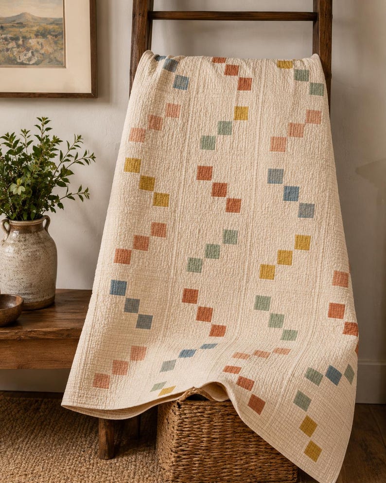 Op de afbeelding: Een cr&egrave;mekleurige quilt met een herhalend patroon van kleine, vierkante lapjes in gedempte tinten blauw, oranje, geel en groen. De stof heeft een zichtbaar getextureerd, doorgestikt oppervlak met verticale stiklijnen door het hele ontwerp.
