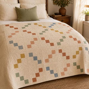 Op de afbeelding: Een cr&egrave;mekleurige quilt met een herhalend geometrisch patroon van kleine, vierkante lapjes in gedempte tinten terracotta, saliegroen, mosterdgeel en stoffig blauw, gerangschikt in diagonale lijnen over het gehele oppervlak van de stof.