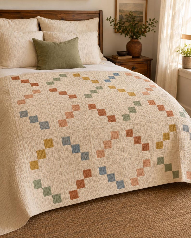 Op de afbeelding: Een cr&egrave;mekleurige quilt met een herhalend geometrisch patroon van kleine, vierkante lapjes in gedempte tinten terracotta, saliegroen, mosterdgeel en stoffig blauw, gerangschikt in diagonale lijnen over het gehele oppervlak van de stof.