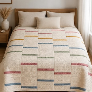 May include: A cream-coloured quilted bedspread featuring a repeating pattern of horizontal stripes in muted shades of dusty rose, mustard yellow, sage green, and slate blue. The quilt has a textured, stitched grid design across the entire surface.