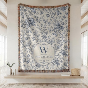 Puede incluir: Una manta con estampado floral azul y blanco con borde con flecos. La manta presenta un emblema central con la letra "W", el apellido "THE WILLIAMS" y el año "2020". La manta se exhibe en una estantería de madera.
