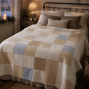 Puede incluir: Una colcha de cama con un patrón de cuadros grandes en cuadrados alternos de color crema, beige y azul claro. La tela tiene una textura acolchada visible en toda su superficie, cubriendo el colchón y cayendo por los lados del marco de la cama.
