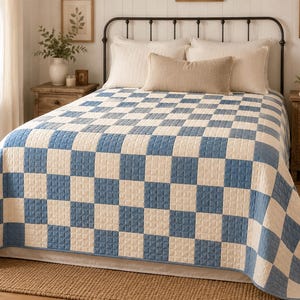 May include: A bed quilt featuring a repeating checkerboard pattern of alternating light blue and off-white square fabric patches. The quilt has a visible quilted texture across its entire surface and covers the top and sides of a bed frame.