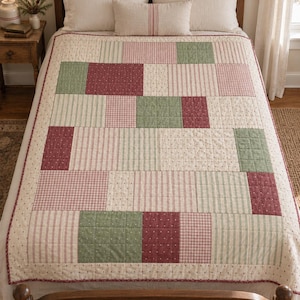 Puede incluir: Una colcha de patchwork con un patrón de bloques rectangulares en tonos verde salvia, rojo oscuro y crema. El diseño incorpora varias texturas de tela, incluyendo cuadros vichy, rayas verticales finas y pequeños estampados florales sobre base crema.