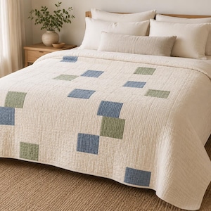 May include: A cream-colored quilt featuring a grid of stitched square patches in muted blue and sage green. The quilt has a textured, quilted surface and covers a bed with matching cream pillows and a long rectangular bolster pillow.