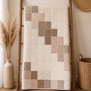 Puede incluir: Una colcha rectangular con un patr&oacute;n de cuadr&iacute;cula de cuadrados en tonos crema, beige, marr&oacute;n claro y rosa suave. La tela tiene una superficie texturizada y acolchada con l&iacute;neas de costura visibles que forman un dise&ntilde;o geom&eacute;trico.
