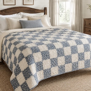 May include: A bed quilt featuring a checkered pattern of alternating solid off-white squares and blue squares with a small floral print. The quilt covers a bed with a dark wood headboard, layered with white and blue pillows, providing a textured surface.