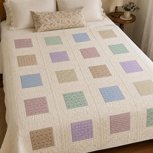 Op de afbeelding: Een crèmekleurige quilt met een raster van vierkante lapjes in diverse pastelkleuren, waaronder lichtblauw, lavendel, saliegroen en beige. Elk vierkant lapje heeft een uniek subtiel patroon en de gehele quilt heeft een doorgestikte textuur.