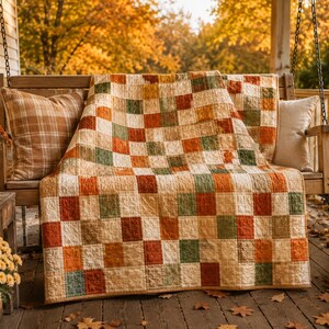 K&ouml;nnte beinhalten: Eine Patchwork-Decke mit einem Gitter aus quadratischen Stoffbl&ouml;cken in den Farben Creme, gebranntes Orange, Olivgr&uuml;n und Braun. Die Decke liegt &uuml;ber einer Holzschaukel, die von zwei Kissen sowie Topf-Chrysanthemen und K&uuml;rbissen umgeben ist.