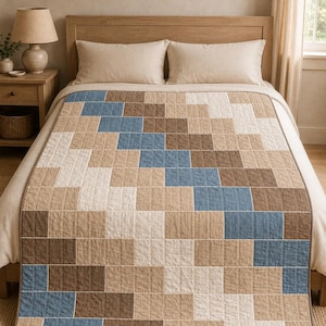 Op de afbeelding: Een rechthoekige quilt met een geometrisch patroon van gestikte stoffen vierkanten in tinten blauw, bruin, beige en gebroken wit. De quilt is gerangschikt in een diagonaal, getrapt ontwerp met zichtbare verticale en horizontale stiklijnen.
