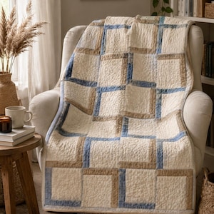 Può includere: Una coperta trapuntata con un motivo geometrico di quadrati color crema, marrone chiaro e blu polvere. Il tessuto presenta un aspetto testurizzato e stropicciato con cuciture visibili, creando un design a blocchi ripetuti su tutta la superficie.