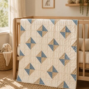 May include: A rectangular quilt featuring a cream-coloured base with a grid of stitched lines. The quilt is decorated with a repeating pattern of squares, each divided into two triangles of muted blue and tan fabric, arranged in a diagonal layout.