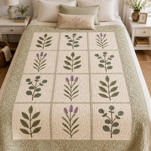 May include: A cream-colored quilt featuring a grid pattern of twelve squares, each containing an embroidered botanical motif of either green leaves or purple lavender sprigs. The quilt has a wide, textured sage green border with a floral pattern.
