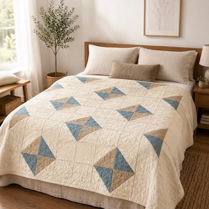 May include: A cream-colored quilt featuring a repeating geometric pattern of squares divided into four triangles. Each square block alternates between solid cream fabric and a combination of two blue and two tan triangles, all with visible stitching.