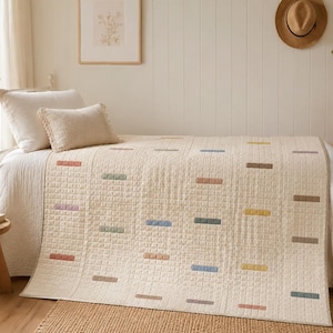 May include: A cream-colored quilt featuring a grid-like texture and small, rectangular embroidered accents in muted shades of pink, blue, yellow, green, and brown, draped over a bed with two matching cream-colored rectangular pillows.