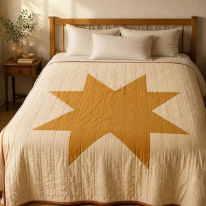 Puede incluir: Una colcha acolchada de color crema que presenta una gran estrella de ocho puntas centrada en tono amarillo mostaza. La colcha tiene líneas de costura verticales y un borde fino de color amarillo mostaza en los extremos, cubriendo una cama.