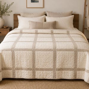 Op de afbeelding: Een crèmekleurige quilt met een ruitpatroon van lichtbeige gestikte lijnen. Het beddengoed ligt over een bed gedrapeerd en toont een gestructureerd stoffen oppervlak met een herhalend vierkant ontwerp dat de hele matras bedekt.