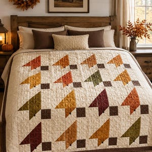 Op de afbeelding: Een crèmekleurige quilt met een herhalend patroon van gestileerde bladvormen in tinten oranje, geel, olijfgroen en bordeauxrood, geaccentueerd met kleine donkerbruine vierkantjes. De quilt heeft een doorgestikte textuur en een donkerbruine rand.