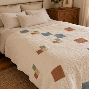 May include: A cream-coloured quilted bedspread featuring a grid pattern of scattered, solid-coloured squares in shades of muted blue, sage green, and terracotta. The quilt has a visible stitched texture and covers a bed with multiple pillows.