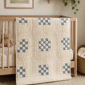May include: A rectangular quilt featuring a cream-colored base with a grid of stitched lines and nine square patches. Each patch contains a blue and cream checkered pattern. The quilt is draped over the side of a light-colored wooden crib.