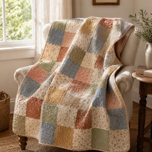 May include: A patchwork quilt in shades of blue, green, and beige, draped over a cream armchair. The quilt is made up of various squares with floral and solid patterns. The chair is placed on a woven rug. The quilt's border is beige.