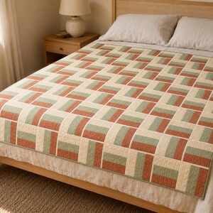 May include: A rectangular quilt featuring a geometric pattern of alternating blocks in muted terracotta orange, sage green, and cream white. The fabric pieces are arranged in a repeating grid layout with visible stitching lines throughout the surface.