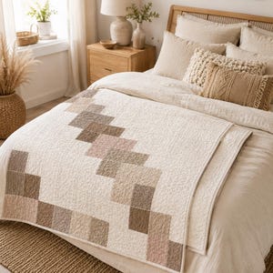 Puede incluir: Una colcha rectangular con una base de color crema y un patr&oacute;n geom&eacute;trico diagonal compuesto por cuadrados cosidos en varios tonos de beige, bronceado, topo y marr&oacute;n suave. La colcha tiene una superficie texturizada y acolchada sobre una cama.