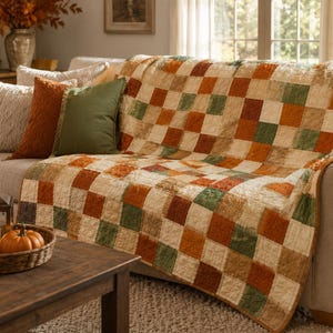 K&ouml;nnte beinhalten: Eine gesteppte Tagesdecke mit einem Karomuster aus abwechselnden Quadraten in Creme, gebranntem Orange und Olivgr&uuml;n. Der Stoff hat eine dezent strukturierte Oberfl&auml;che und die Decke liegt &uuml;ber der R&uuml;ckenlehne und Sitzfl&auml;che eines neutralen Sofas.