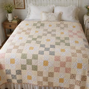 Puede incluir: Una colcha de patchwork con un patrón de cuadrícula repetitivo de cuadrados en tonos suaves de crema, verde salvia, rosa empolvado, amarillo mostaza y gris apagado. El diseño incorpora bloques de tela con estampado floral y lisos.