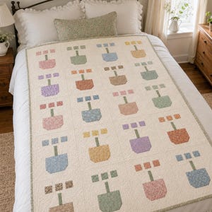 May include: A rectangular quilt featuring a repeating grid pattern of potted flower motifs in various pastel colours including pink, blue, yellow, green, and brown. The quilt has an off-white background and a thin green floral border trim.