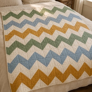 Peut inclure: Une courtepointe rectangulaire présentant un motif de chevrons répétitif dans des tons sourds de vert sauge, bleu poussiéreux et jaune moutarde, séparés par des bandes de tissu crème. La courtepointe présente des coutures en grille visibles.