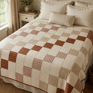 May include: A bed quilt featuring a patchwork design of square blocks in shades of cream, beige, dusty rose, and terracotta. The quilt has a visible grid-like quilted texture and a solid terracotta-colored fabric border along the bottom edge.
