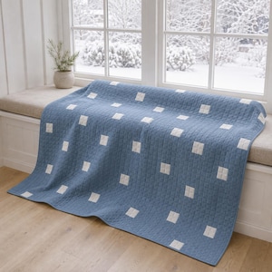 May include: A rectangular quilt featuring a dusty blue base color with a grid of small, white square accents distributed across the surface. The fabric has a visible stitched texture throughout, creating a uniform pattern of squares.