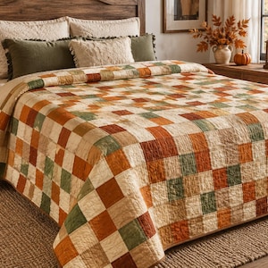 K&ouml;nnte beinhalten: Eine Bettdecke mit einem sich wiederholenden Patchwork-Muster aus quadratischen Stoffbl&ouml;cken in den Farben gebranntes Orange, Olivgr&uuml;n und Creme. Die Decke liegt auf einem Bett und zeigt ein durchgehendes Rasterdesign mit sichtbaren N&auml;hten.