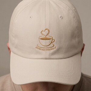 May include: A beige baseball cap featuring an embroidered gold-coloured design of a coffee cup with a heart-shaped steam swirl above it. Below the cup, the text "ESPRESSO YOURSELF" is stitched in gold thread, centred on the front panel.