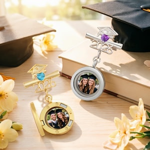 Puede incluir: Dos medallones de fotos con temática de graduación, uno dorado y otro plateado, cada uno con una foto pequeña en su interior. El medallón dorado está abierto, revelando una foto. Ambos medallones tienen un diseño de birrete y pergamino con el año 2026.