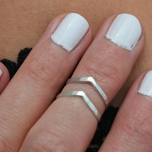 May include: A close-up of a hand wearing two silver chevron rings on the middle finger. The fingernails are painted white.