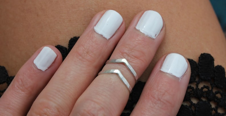 May include: A close-up of a hand wearing two silver chevron rings on the middle finger. The fingernails are painted white.