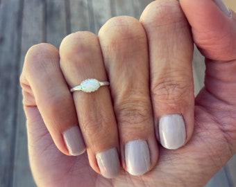 May include: A silver ring with a white oval opal gemstone. The ring is on a finger with light pink nail polish.