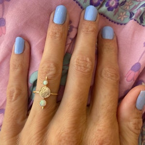 May include: A gold ring featuring a textured circular design with two small, round, white opal stones and a vertical bar with a third opal. The ring is worn on a hand with light blue nail polish.