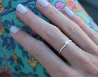 May include: A thin silver ring featuring a small, round, white opal stone set in a bezel. The band of the ring has a textured, hammered finish that catches the light, providing a subtle, reflective surface across the entire circumference of the band.