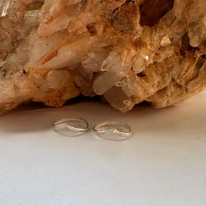 May include: Two silver rings with a curved, open design. The rings are on a white surface in front of a large, light brown rock.