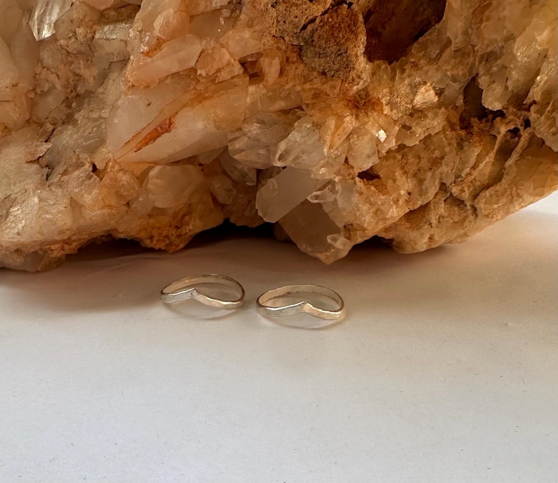May include: Two silver rings with a curved, open design. The rings are on a white surface in front of a large, light brown rock.